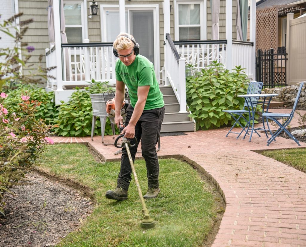 Man weed wacking a customers grass next to a sidewalk with the house in the background.